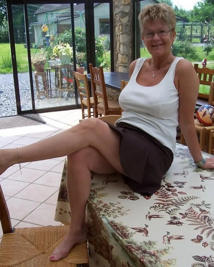 Femme aux cheveux blonds courts, portant un haut blanc, une jupe marron et des sandales, assise sur une table en extérieur, une jambe posée sur une chaise.