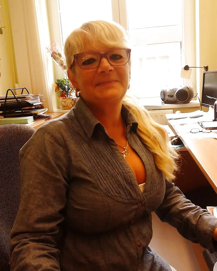 Femme aux cheveux blonds courts, portant une chemise grise boutonnée, assise à un bureau devant une fenêtre, souriant légèrement.