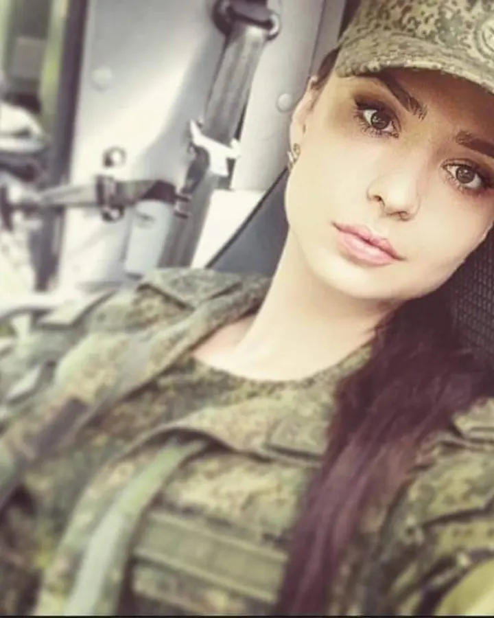 Femme en tenue militaire camouflée, casquette, regardant l'appareil photo depuis l'intérieur d'un véhicule.