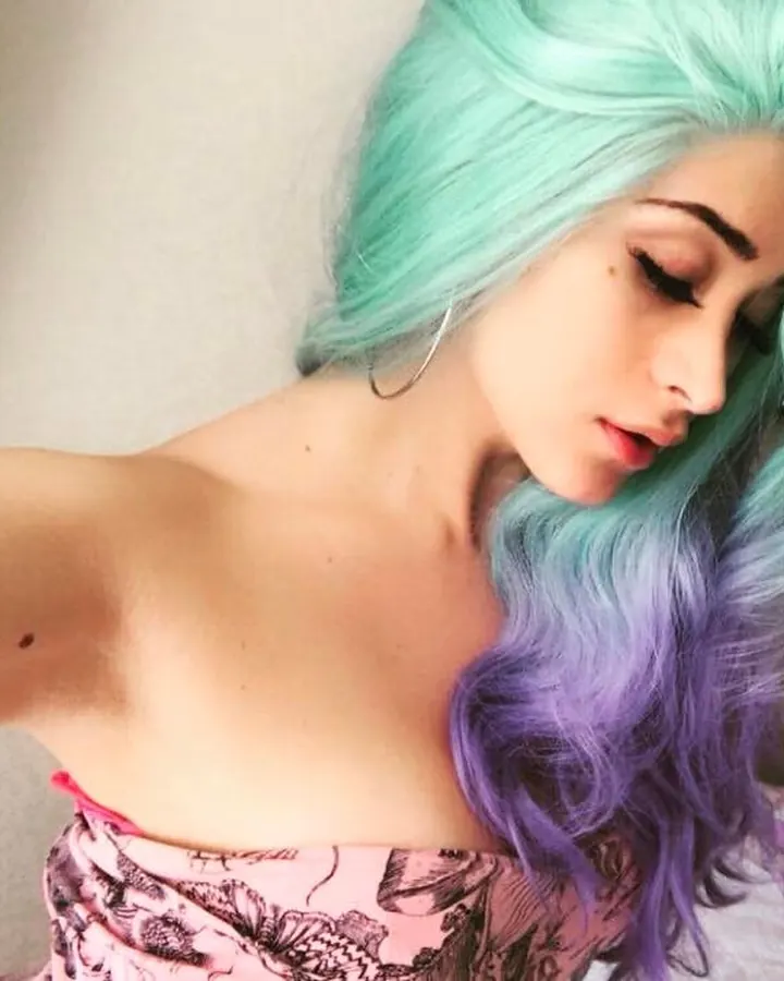 Jeune femme aux cheveux bruns-noirs, portant une perruque turquoise et violette, prenant un selfie dans une chambre, avec un haut rose et un téléviseur en arrière-plan.