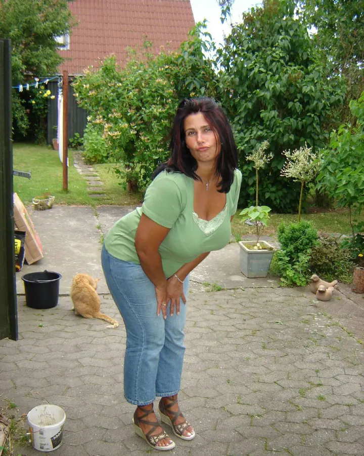 Femme aux cheveux bruns, portant un haut vert et un jean bleu, posant dans un jardin, avec un chat orange en arrière-plan.