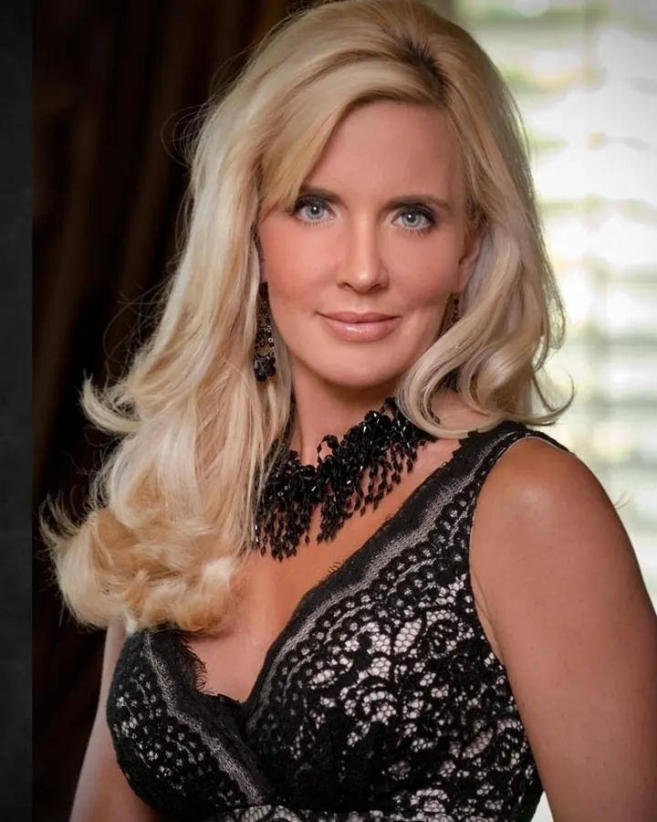 Femme blonde aux yeux bleus, portant une robe noire en dentelle, posant dans un intérieur sombre, avec des boucles d'oreilles pendantes.
