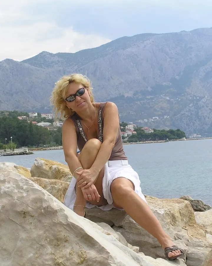 Femme blonde aux cheveux courts, portant un haut marron et une jupe blanche, assise sur des rochers près d'une mer, avec des montagnes en arrière-plan.