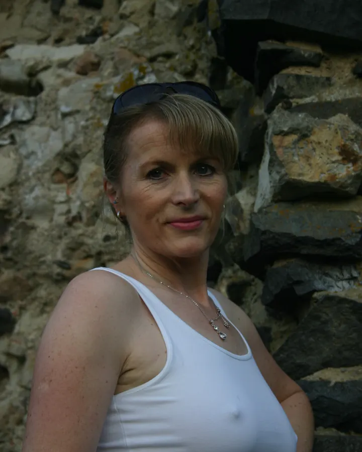 Femme européenne aux cheveux blonds, portant un débardeur blanc, posant devant un mur en pierre, avec des lunettes de soleil sur la tête.
