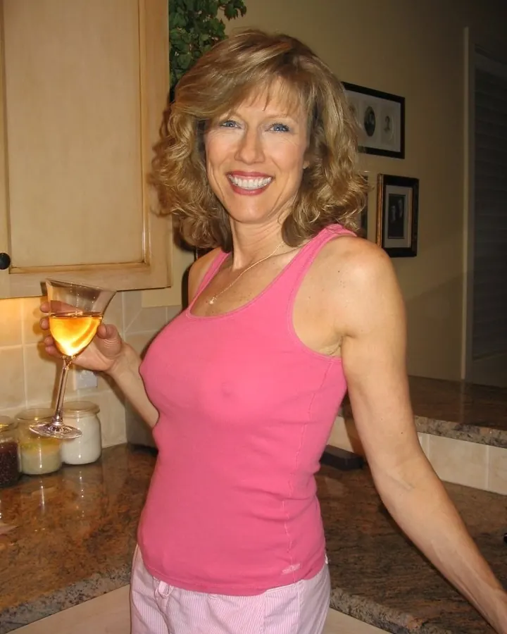 Femme blonde en débardeur rose, tenant un verre de vin, souriant dans une cuisine.