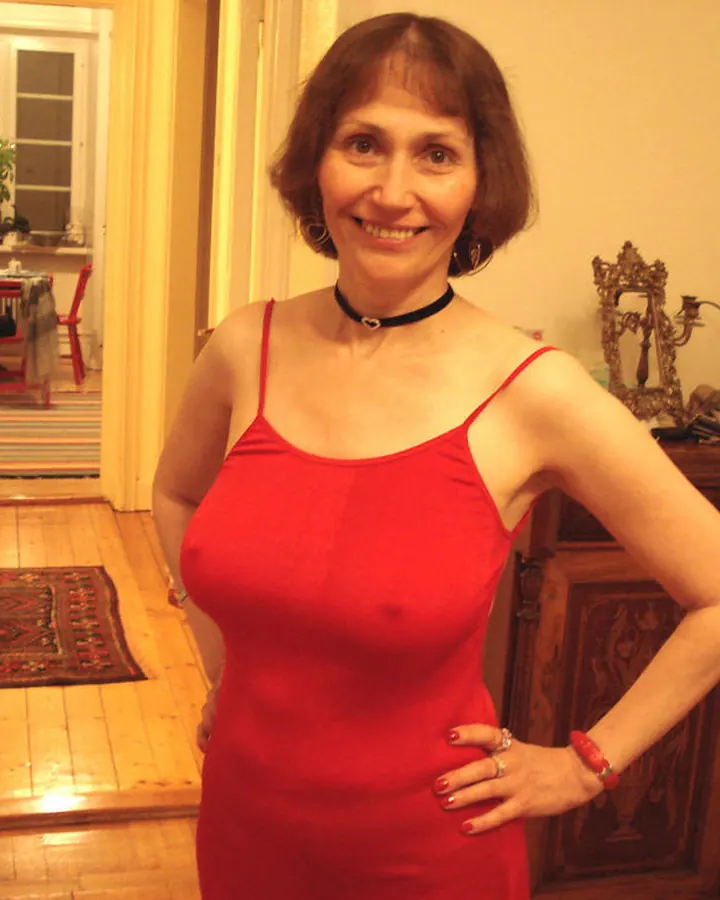 Femme aux cheveux châtains en robe rouge, souriant, debout dans un couloir avec un meuble en bois derrière elle.