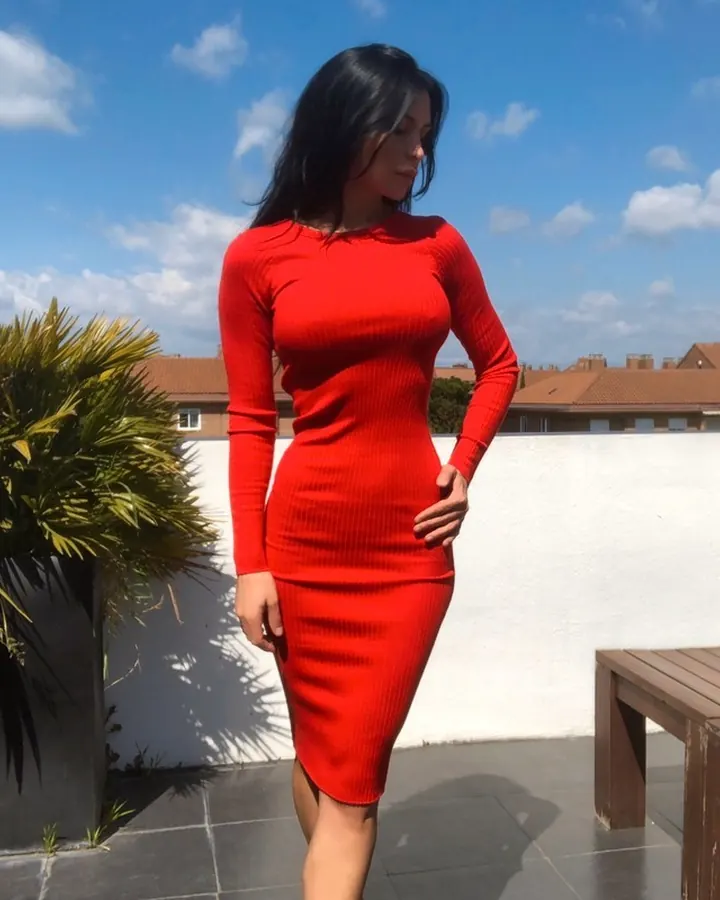 Femme aux cheveux longs, vêtue d’un haut noir à rayures blanches, penchée sur un buisson, portant des lunettes de soleil et des chaussures à talons noirs et blancs