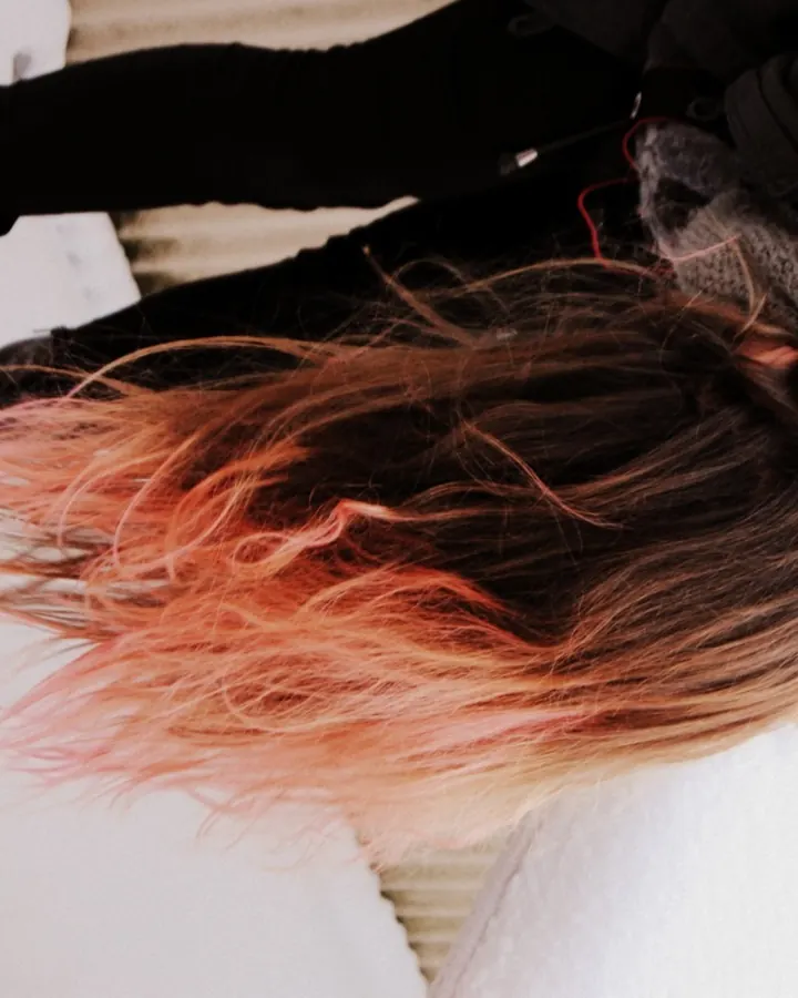 Femme allongée sur la neige, vue de dessus, cheveux longs aux pointes roses, vêtue de noir, avec un foulard gris.