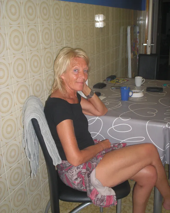 Femme blonde aux lunettes, en chemisier noir, souriant, avec des lunettes de soleil sur la tête.