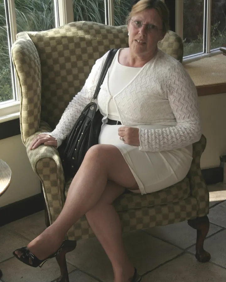Femme blonde aux lunettes, en chemisier blanc et jupe blanche, assise dans un fauteuil à carreaux.