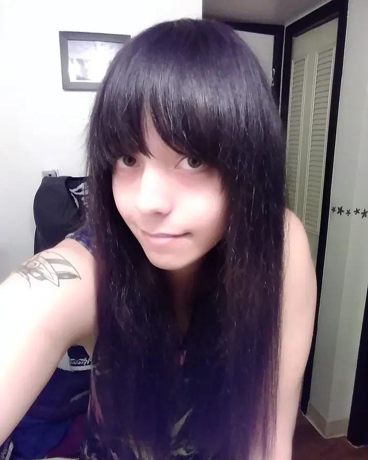 Jeune femme aux cheveux noirs longs, regardant l'appareil, tatouage visible sur le bras gauche, dans une pièce intérieure.