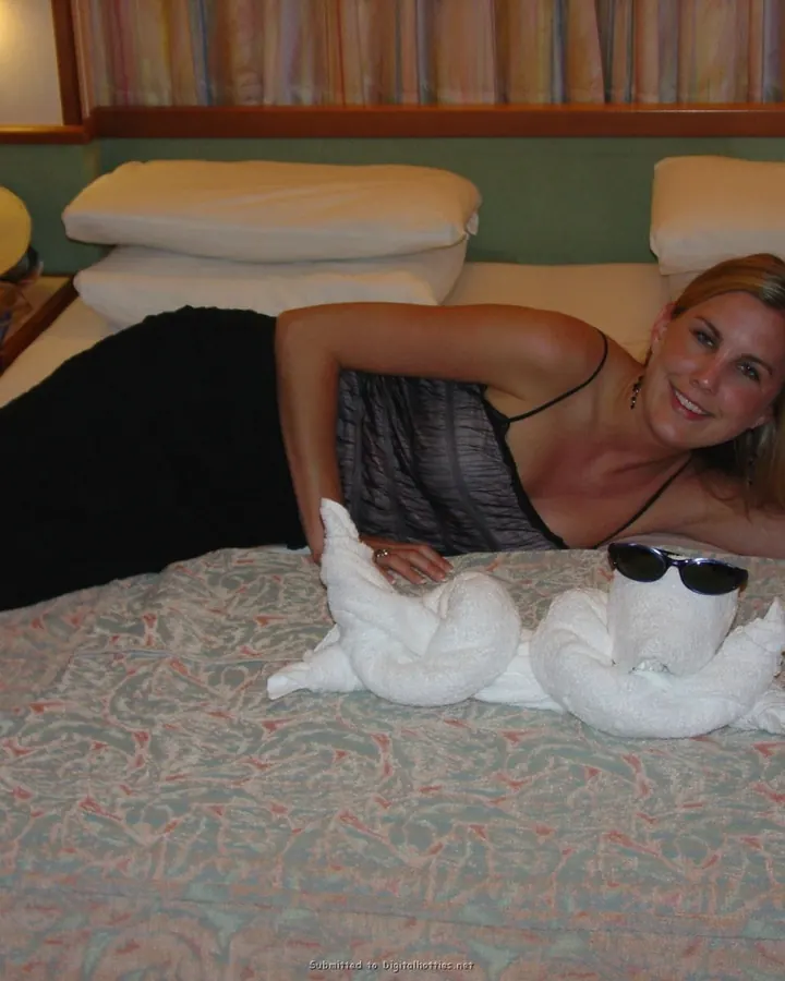 Femme blonde, allongée sur un lit dans une chambre, en tenue noire, souriant à l'appareil, avec deux serviettes pliées en forme d'animaux devant elle.