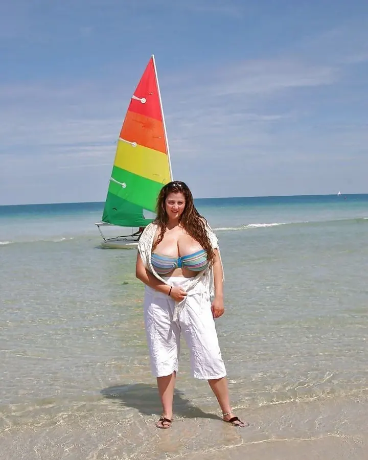 Femme aux cheveux châtains, seins volumineux, en maillot rayé bleu et blanc, debout dans l'eau claire d'une plage, avec un voilier coloré en arrière-plan.