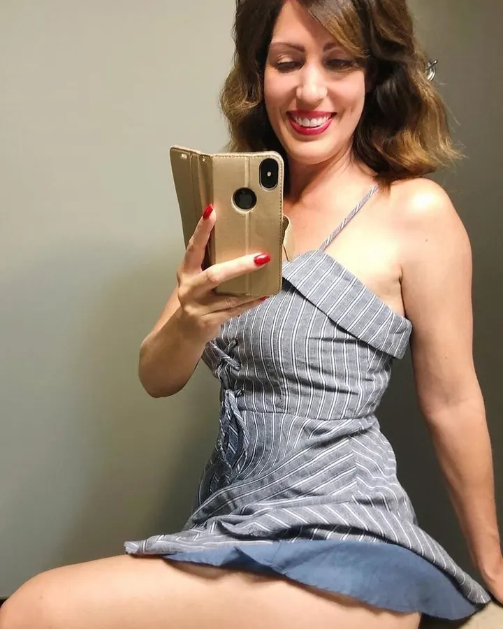 Femme aux cheveux bruns, souriante, prenant un selfie avec un téléphone doré, vêtue d'une robe grise à fines rayures.
