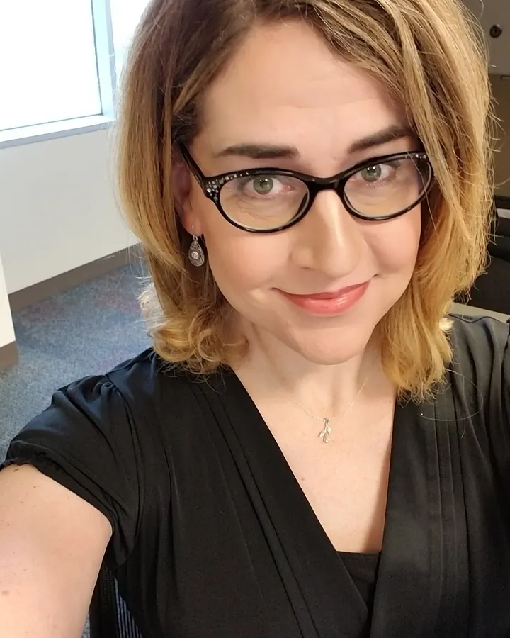Femme aux cheveux blonds courts, portant des lunettes, souriant, vêtue d'une robe noire, en selfie dans un bureau.