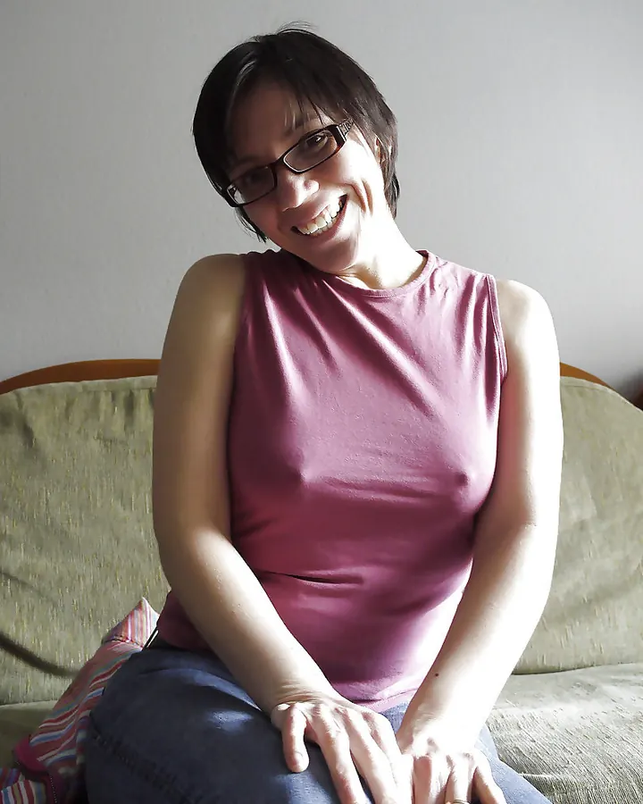 Femme aux cheveux bruns courts, portant un débardeur rose, souriante, assise sur un canapé, en intérieur.