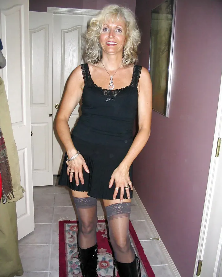 Femme blonde en robe noire, debout dans un couloir, portant des bottes noires et des collants à motifs